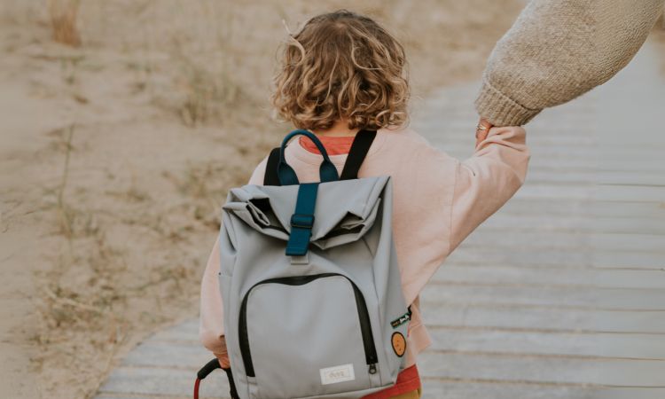 Kind spaziert am Strand mit Mutter an der Hand und den nachhaltigen Rucksack von Dusq am Rücken - Kinderrucksack aus recyceltem Material, fair hergestellt in Europa, mit reflektierenden Applikationen